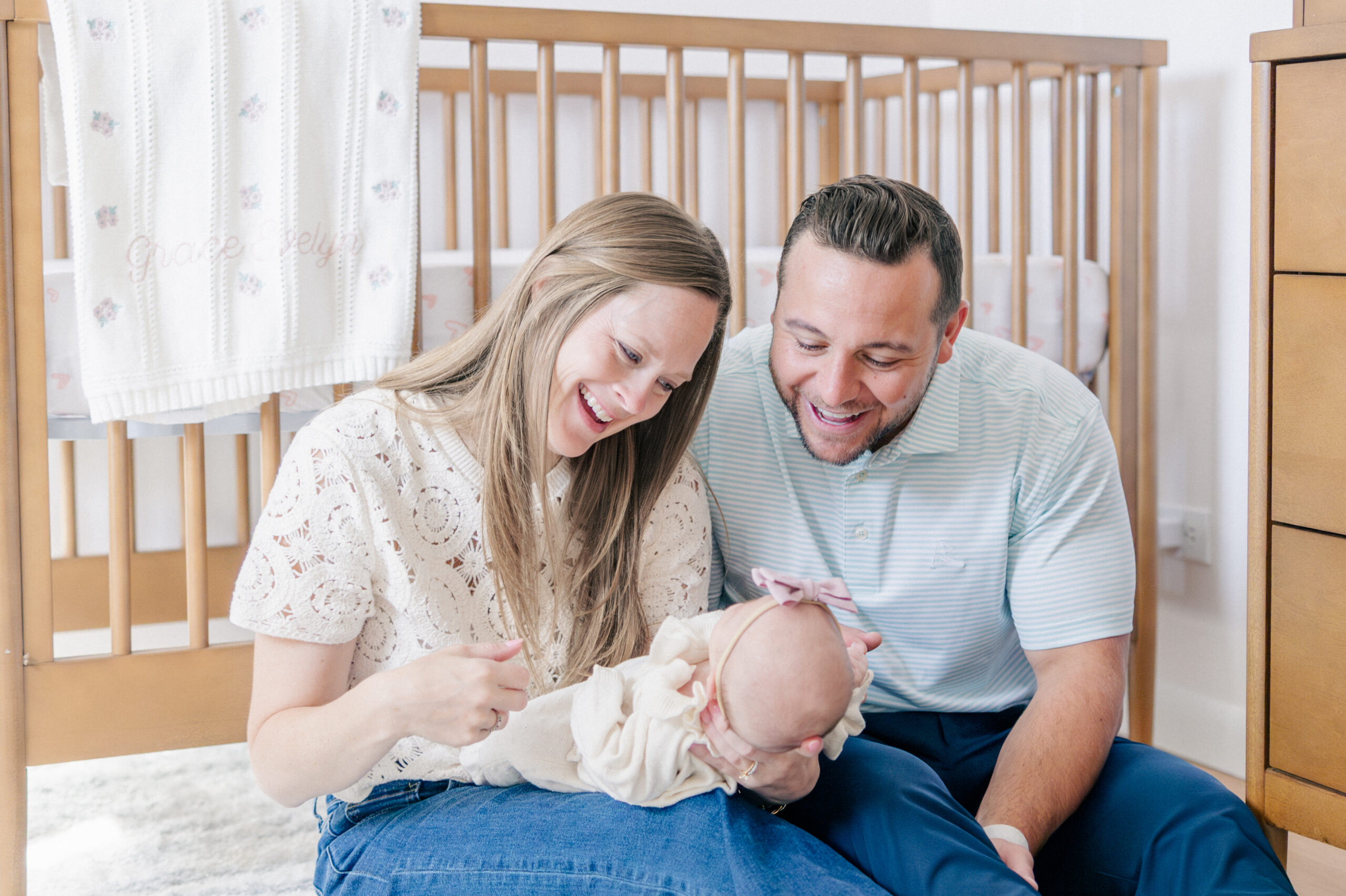 Lifestyle newborn photo of baby sleeping in parents’ arms next to crib with natural light
